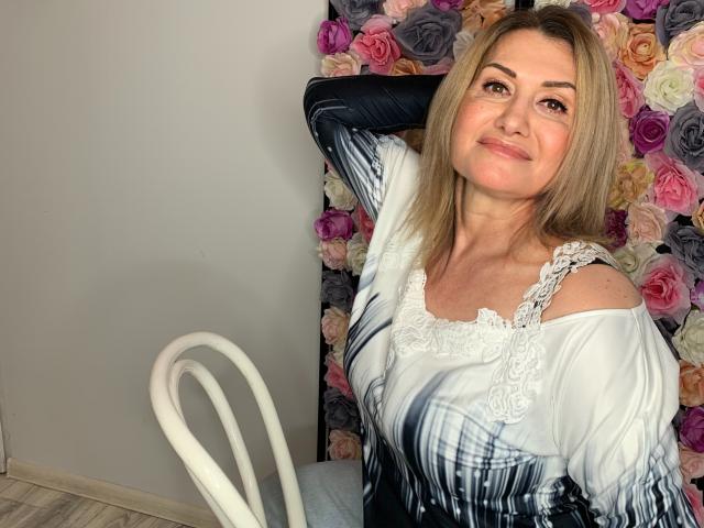 IsabellaSensual, a middle-aged woman with blonde hair, smiling, has her arm raised, wearing a white dress with black and gray patterns, sitting in front of a floral wall.