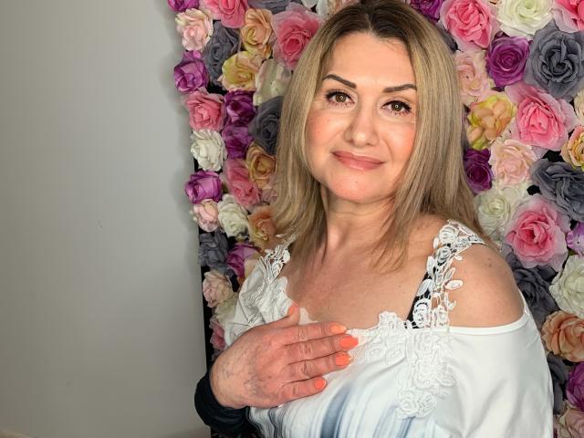 IsabellaSensual with blonde hair, light skin, and small tits, wearing a white lace top, smiling, hand on chest, pink and purple roses in background, neon orange nails.