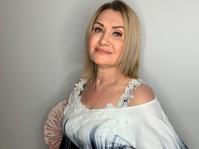 IsabellaSensual has blonde hair, fair skin, and a gentle smile. She's wearing a lace-off shoulder top with white and blue gradient, holding a pink fan.