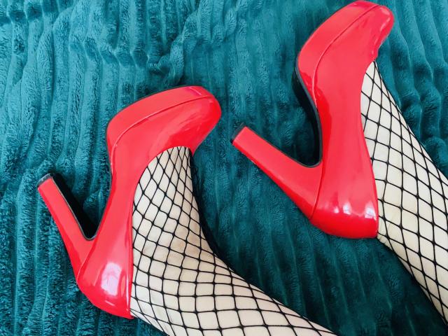 KarKutie's shiny red high heels with black fishnet stockings, her legs are spread apart, showing the heels' pointed toes and stiletto heels.