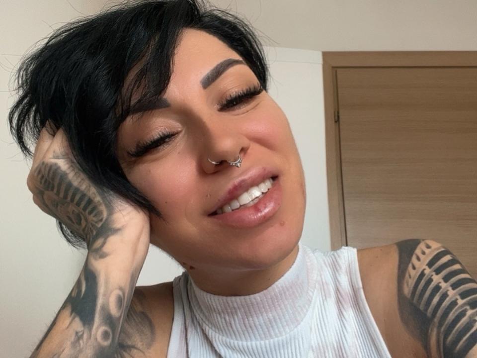 JudithRuhh with short black hair, nose piercing, tattoos on arms, white sleeveless top, smiling, eyes closed, hand in hair, shiny pink lips, light skin, wooden door in background.