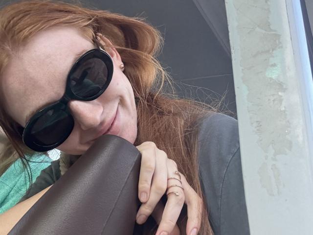 GingerFairy with red hair, wearing black sunglasses, smiling, resting head on brown leather, hands with rings, gray shirt, and green towel in background.