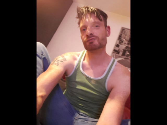 Defender-hot has a green tank top, visible tattoo on his right arm, and is sitting with a slight smirk. He's wearing blue jeans and has a short, messy haircut.