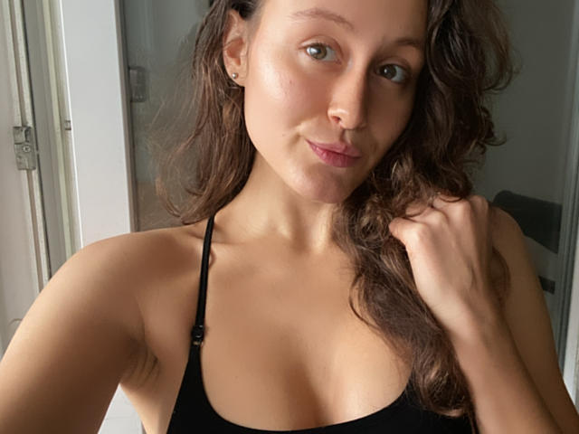 AlesyaDrons with wavy brown hair, light skin, small tits, wearing a black halter top, smiling softly, holding her hair with her right hand, standing near a window.