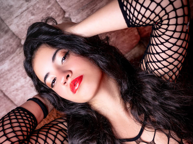 NicolSky with red lips, black fishnet sleeves, and wavy dark hair, lying down with arms behind head, looking up with sultry expression, showing off her tits and pussy.
