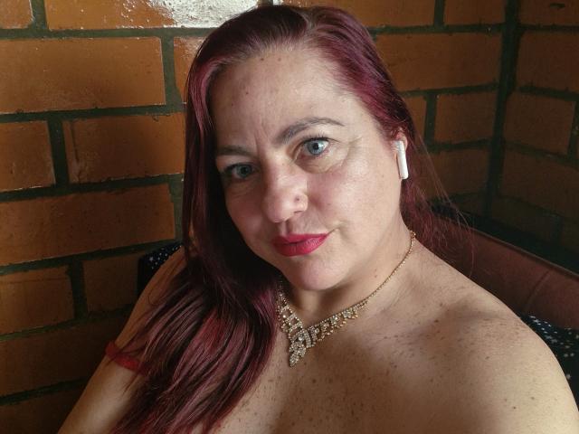 LovelyScarlett with red hair, freckled skin, and red lipstick, wearing a jeweled necklace, has her tits exposed, and is sitting against a brick wall.