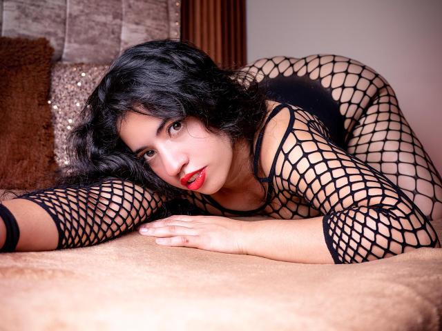 NicolSky with black fishnet lingerie, red lipstick, and curly hair, on a bed, face down, looking at camera, ass up, hands on bed, sensual expression, sexy pose.