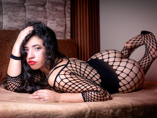NicolSky lies on a bed, wearing a black fishnet bodysuit, exposing her ass and pussy. She has long black hair, red lipstick, and a sultry expression.