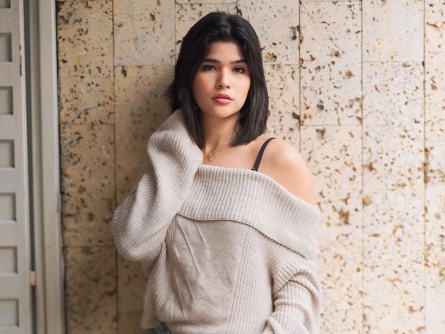 ScarlletZhen with black shoulder-length hair, off-shoulder beige sweater, black strap, subtle makeup, standing against a speckled wall, looking at camera, hands near chest.