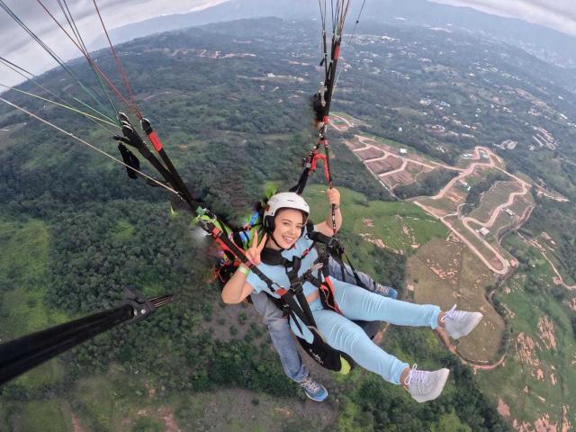 ZoeTobon69 with a happy smile, wearing a white helmet, blue jeans, and white sneakers, hanging from a zipline with multiple ropes, green forest below, and a clear sky.