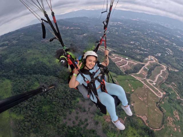 ZoeTobon69, with a white helmet, blue leggings, and white sneakers, is hanging from a zip line, smiling, with multiple black and red cables attached to her.