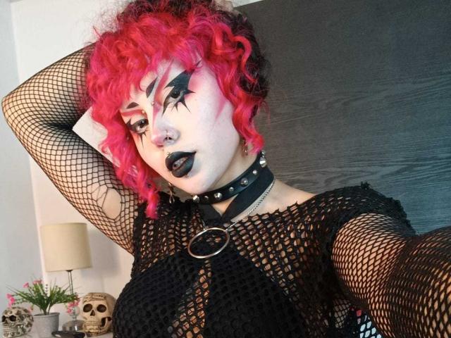 GothVioleth met rood krullend haar, een zwarte fishnet top, een choker met spikes, zwarte ster-make-up en donkere lipstick, die haar borsten laat zien, met een zwarte ring met studs in haar borst..