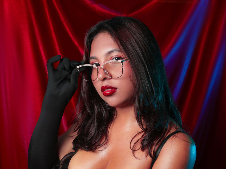 SashaConnor with glasses, red lipstick, black gloves, and medium tits, holding glasses, against red and blue fabric. She's got long brown hair and a sexy, confident look.