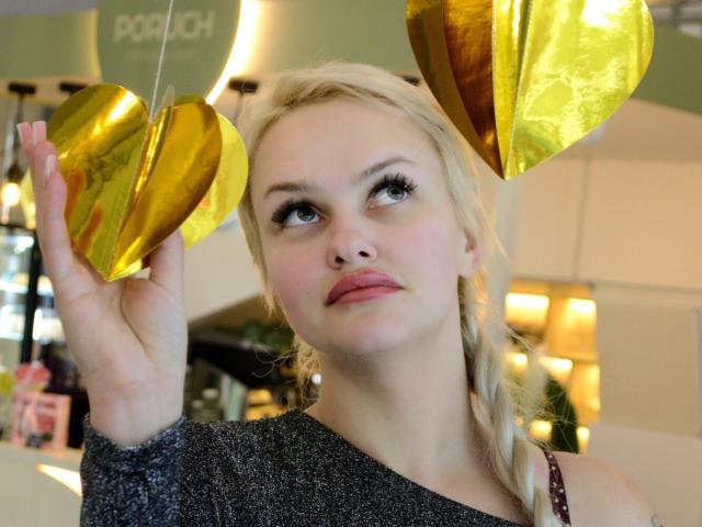 Melissa, une femme aux cheveux blonds, tenant des seins dorés brillants, regardant vers le haut, portant un haut gris scintillant, dans un environnement intérieur bien éclairé. Elle a une expression pensive..