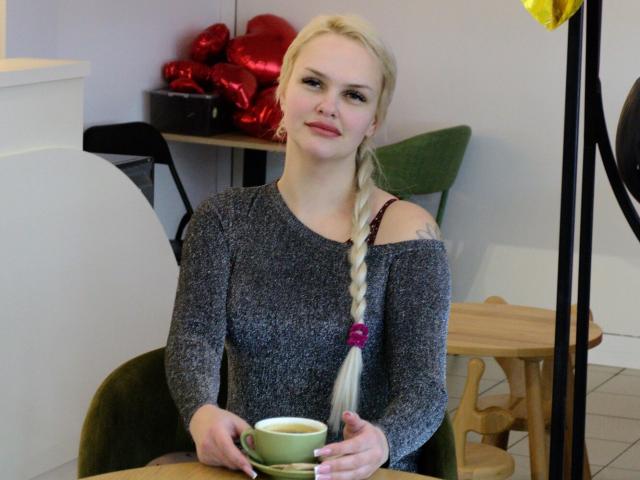 Melissa: Kat met blond vlechtje, grijze sweater met open schouder, vasthoudend een groene beker, zittend in een café, lachend, roze haarbandje, kleine tatoeage op de rechter schouder, houten tafel..