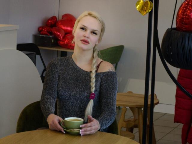 Melissa, eine Katze mit blondem Zopf, trägt einen grauen Pullover mit offenem Ausschnitt, hält eine grüne Tasse und sitzt an einem Holztisch. Im Hintergrund sind rote und gelbe Luftballons zu sehen, auf der rechten Seite hängt ein schwarzes Ornament..