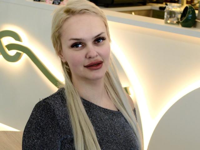 Melissa: Gatas com cabelos loiros longos, vestindo um suéter cinza, sorrindo levemente, em pé em uma área interna bem iluminada, com iluminação suave e decoração verde..
