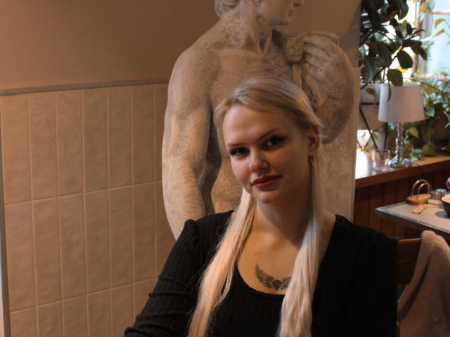 Melissa: Een vrouw met blonde vlechtjes, een zwart gestreept shirt en een tatoeage onder haar kin, die voor een hoge stenen sculptuur staat waarbij haar borsten licht zichtbaar zijn, en die glimlachend naar de camera kijkt..