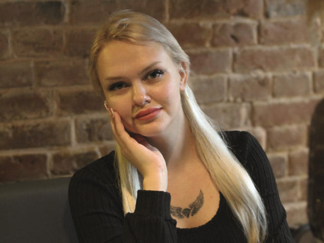 Melissa, eine Katze mit blonden Haaren, einem schwarzen Pullover, einem Nasenpiercing und einem Flügel-Tattoo auf der Brust, die ihren Kopf auf ihre Hand legt und sanft lächelt..