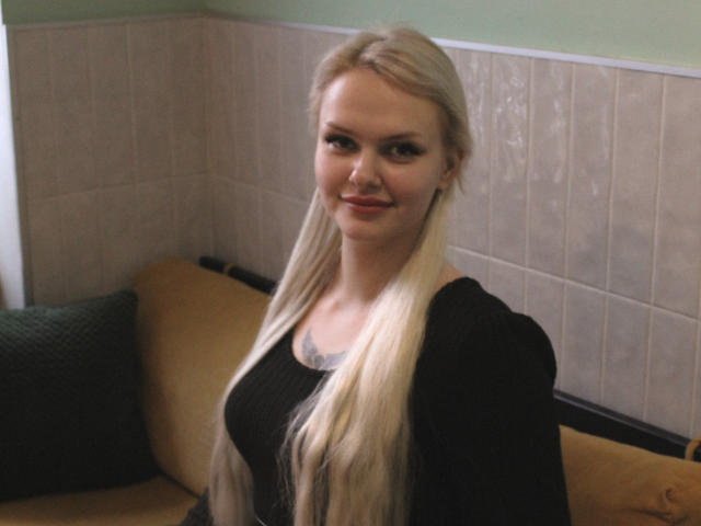 MelissaCats hat langes, blondes Haar, ein schwarzes Oberteil und ein Tattoo unter ihrer Brust. Sie lächelt und hat kleine Brüste, grüne Augen und helle Haut. Sie sitzt auf einem gelben Sofa..