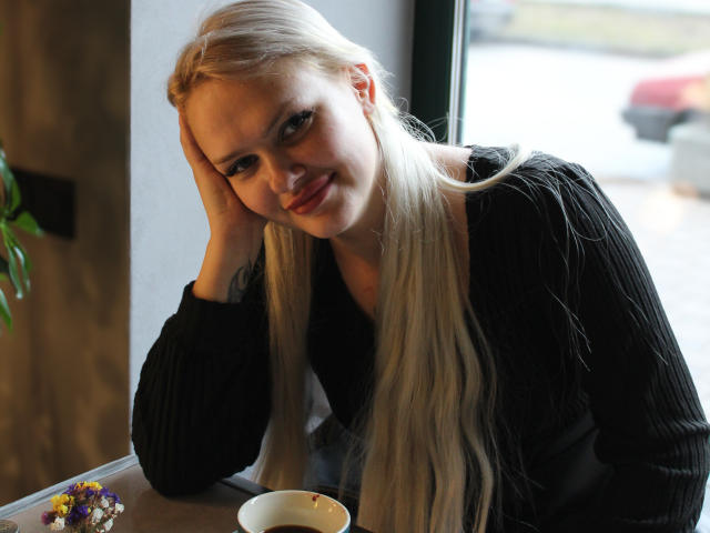 Melissa, eine Katze mit langem, blondem Haar, einem schwarzen, gerippten Pullover und einem süßen Lächeln, ruht ihren Kopf in ihrer Hand, neben einer Schüssel und einem bunten Snack, in einem gemütlichen Café..