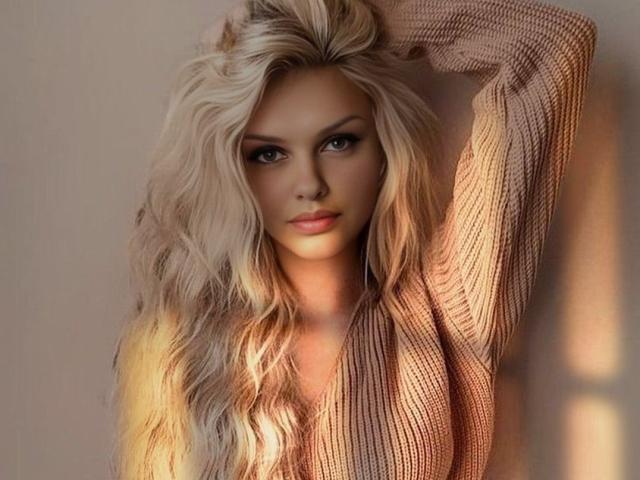 MelissaCats hat langes, welliges blondes Haar und trägt einen beige-farbenen, gerippten Pullover mit langen Ärmeln. Ein Arm ist angehoben, und sie blickt direkt in die Kamera mit einem neutralen Gesichtsausdruck..