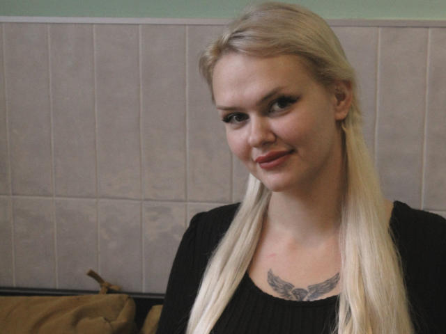 MelissaCats tem cabelo loiro, blusa preta e uma tatuagem no peito com um desenho de asas. Ela está sorrindo, com pele clara e maquiagem discreta, e está em frente a uma parede de azulejos..