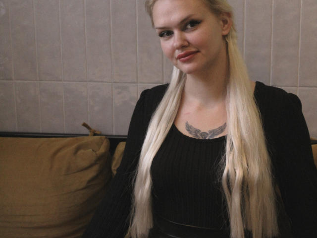 Eine Melissa, mit langen blonden Zöpfen, einem schwarzen, gerippten Oberteil, einem kleinen Brusttattoo und einem leichten Lächeln, sitzt auf einem braunen Kissen..