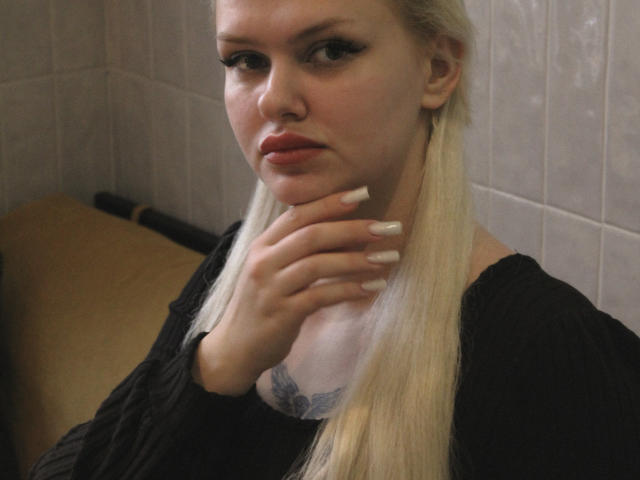 Melissa: Een vrouw met lang blond haar, een zwarte top en witte nagels, die haar nek aanraakt en een tatoeage onder haar kin laat zien, kijkend naar de camera met een serieuze uitdrukking..