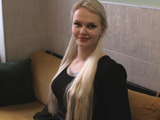 MelissaCats heeft lang, steil blond haar, kleine borsten en een zwarte top, en zit op een gele bank met een groen kussen, waarbij ze zachtjes naar de camera glimlacht..