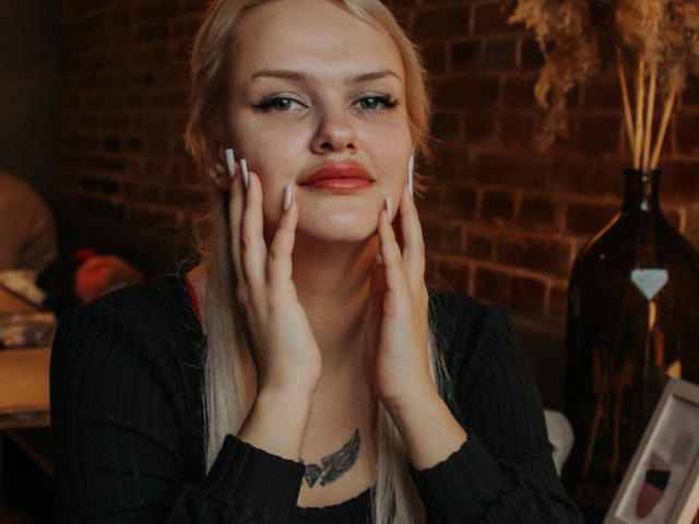 Melissa. Donne con capelli biondi, mani sulle guance, camicia a righe nere, rossetto rosso, unghie bianche, tatuaggio ad ali sul petto, muro di mattoni, vaso con piante secche sullo sfondo..