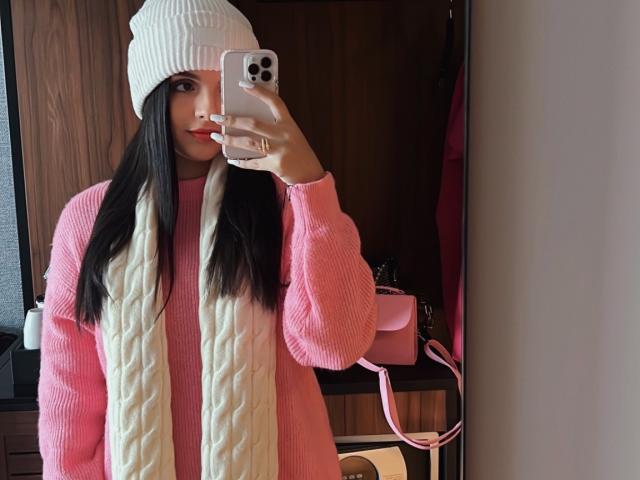 NovaDixon with long dark hair, wearing a white beanie, pink sweater, and cream scarf, taking a mirror selfie with an iPhone. Pink purse and wooden wall in background.