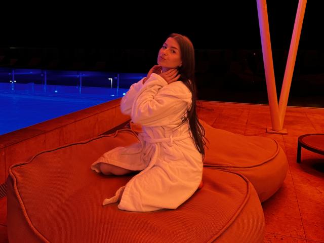 AnnettMonro with long brown hair, kneeling on orange cushions, wearing a white robe, hands near her chin, by a blue-lit pool at night, in a dimly lit area.
