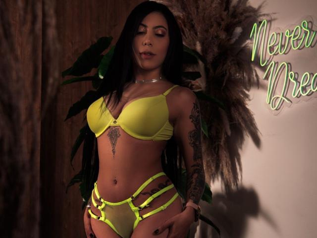 AmeliaRuso with long black hair, yellow lingerie, and tattoos, standing, showing her small yellow bra and strappy yellow panties, with her hands on her hips, looking down.