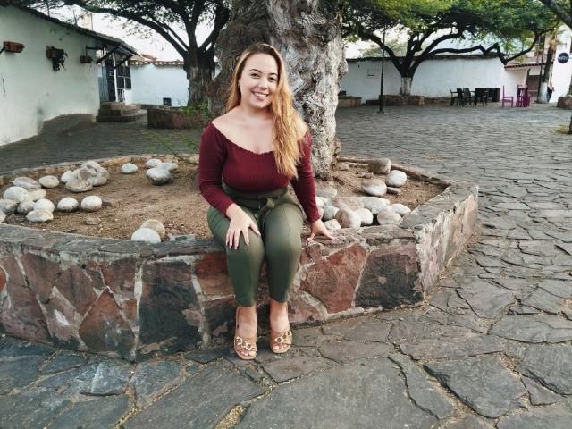 ZoeTobon69, with long blonde hair, sits on a stone bench, wearing a red top, green pants, and sandals, smiling, with a tree and rocks behind her.