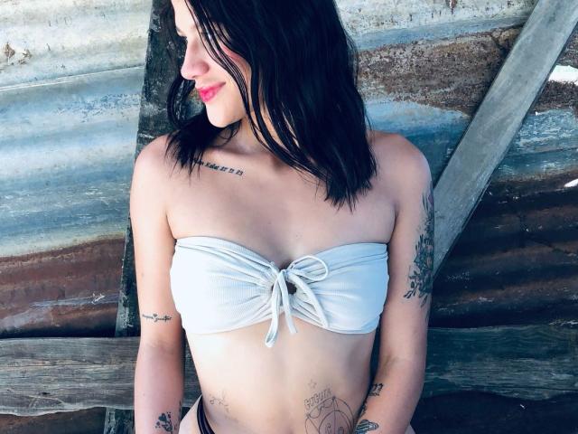 Chelsey Soto, con i capelli neri, indossa un top di bikini bianco senza spalline, che mette in mostra piccoli seni, ha tatuaggi sulle braccia e sul petto, sorride con il rossetto rosso e sta in piedi contro un muro di legno..