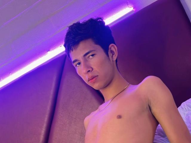 MatthSexy, shirtless, with a lean build, small nipples, and short dark curly hair, looks off to the side with a slight pout, under purple LED lighting.
