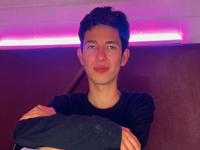 MatthSexy, with short dark hair and a slight smile, crosses his arms in a black long-sleeve shirt, under pink neon lighting. He's sitting on a brown leather seat.