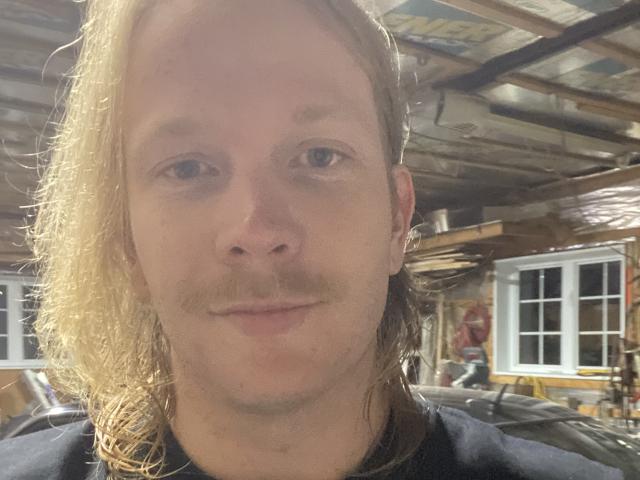 WayneLust with long blonde hair, mustache, and a slight smile, wearing a black shirt, has a fair complexion and blue eyes in a dimly lit room.