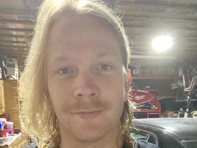 WayneLust has blonde, shoulder-length hair, a light mustache, and a slight smile. His blue eyes are centered in the image. He's in a dimly lit garage.