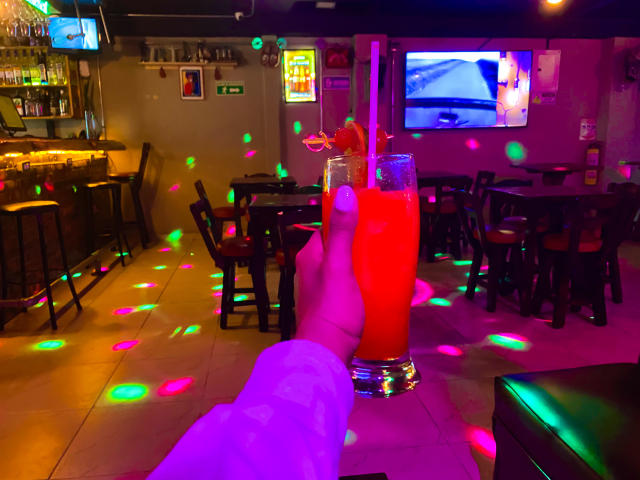 SophieSonyas' hand holding a tall, bright red cocktail with a cherry and lemon slice, surrounded by colorful laser lights in a dimly lit bar.