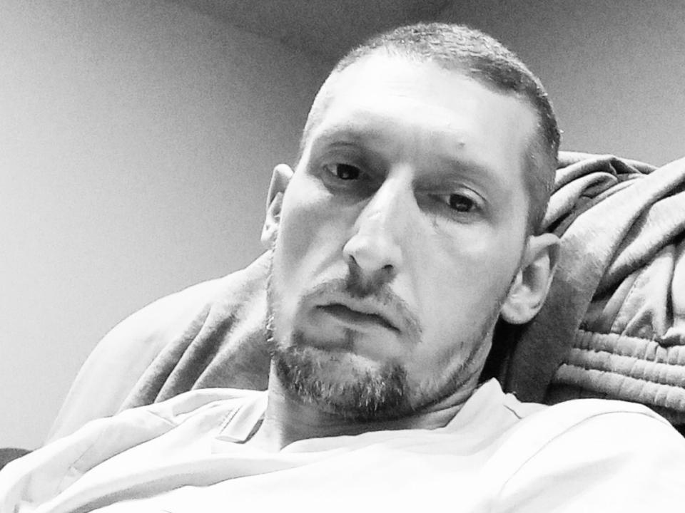 MarkDzara with a serious face, short buzzcut, light stubble, and a slight beard, wearing a light shirt, lying down with a blanket behind him. Black and white photo.