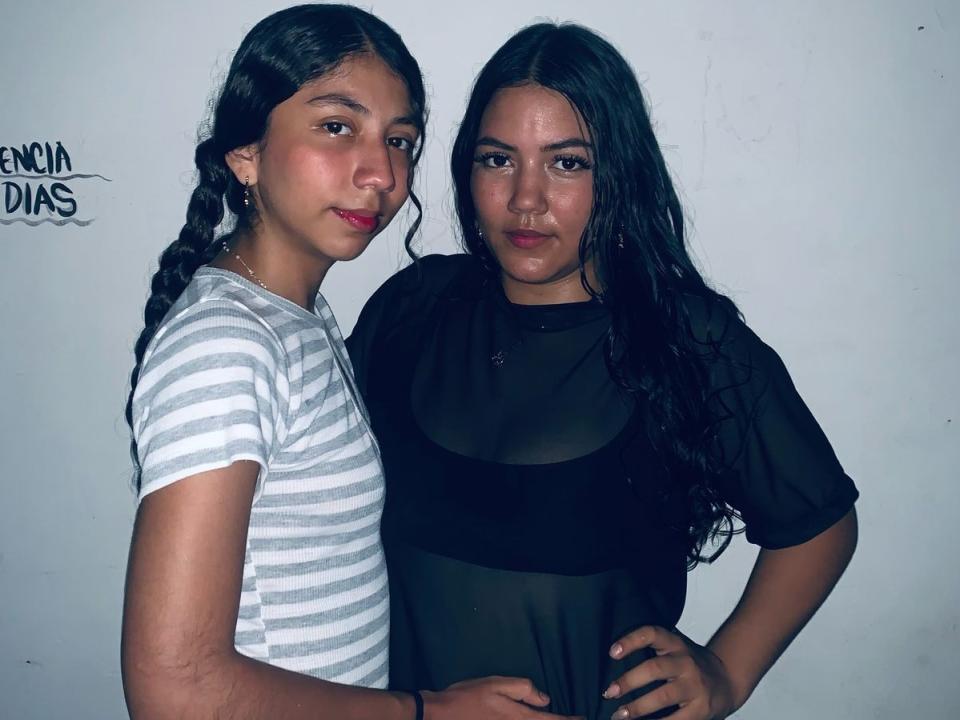 LolaAndJenny with dark hair, one in a striped shirt, the other in a black top, standing side-by-side, hands on hips, both looking at the camera.