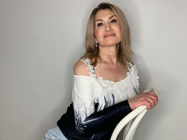 IsabellaSensual with blonde hair, off-shoulder white lace top, black sleeves, sitting on white chair, smiling slightly, holding chair, light skin, small earrings, relaxed, attractive.