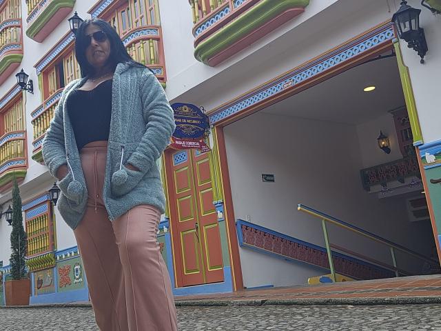 JackieMature, black woman, long hair, wearing black top, pink pants, blue fuzzy cardigan, standing in front of colorful building, hands in pockets, wearing sunglasses, street lamp in background.