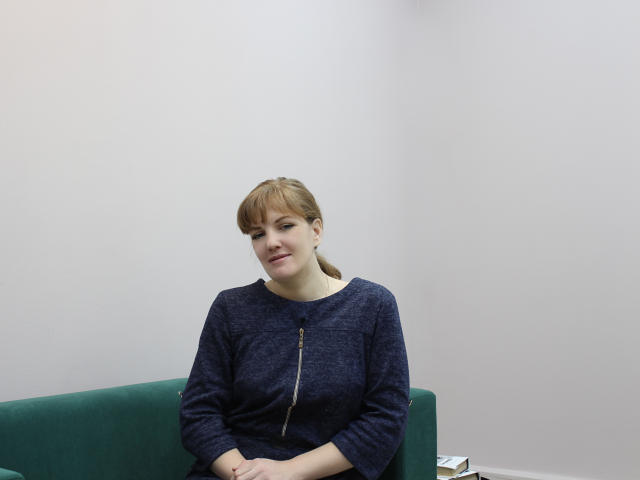 AlissaAmazing has light skin, brown hair in a ponytail, and wears a blue sweater with a zipper. She's sitting on a green couch, smiling slightly.