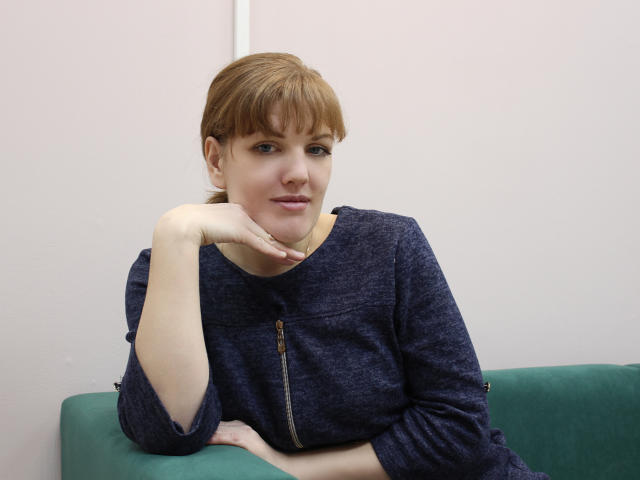 AlissaAmazing, with red hair and bangs, wears a navy zip-up sweater, leaning on a green couch, her hand under her chin, smiling slightly, looking relaxed.