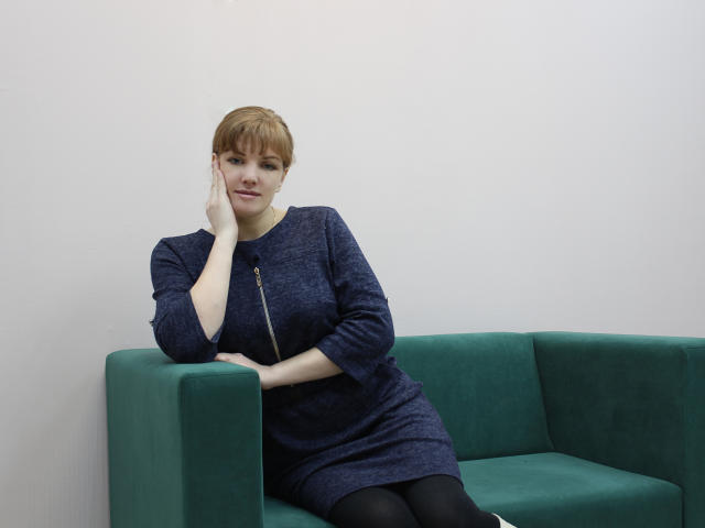 AlissaAmazing with short red hair, wearing a blue dress and black tights, sits on a green couch, hand on cheek, looking slightly to the side, smiling subtly.