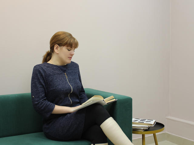 AlissaAmazing, a red-haired chick with a zipper dress, black leggings, and white boots, reads on a green couch, next to a small yellow table with magazines.