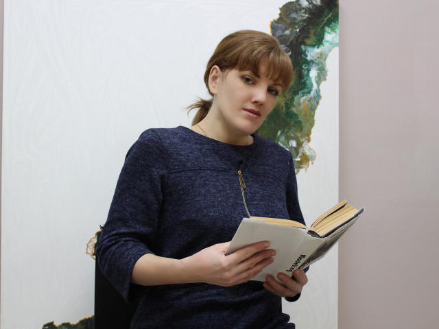 AlissaAmazing with brown hair in a ponytail, wearing a navy blue sweater, holding an open book, looking slightly to the side, standing in a well-lit room.